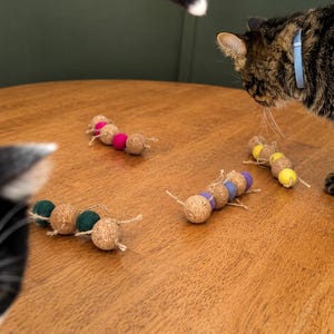 May include: Several cat toys made of cork and colorful felt balls are arranged on a wooden table. The toys are in a caterpillar-like design, with each segment a cork ball and a felt ball. A cat's tail and two cats are visible.