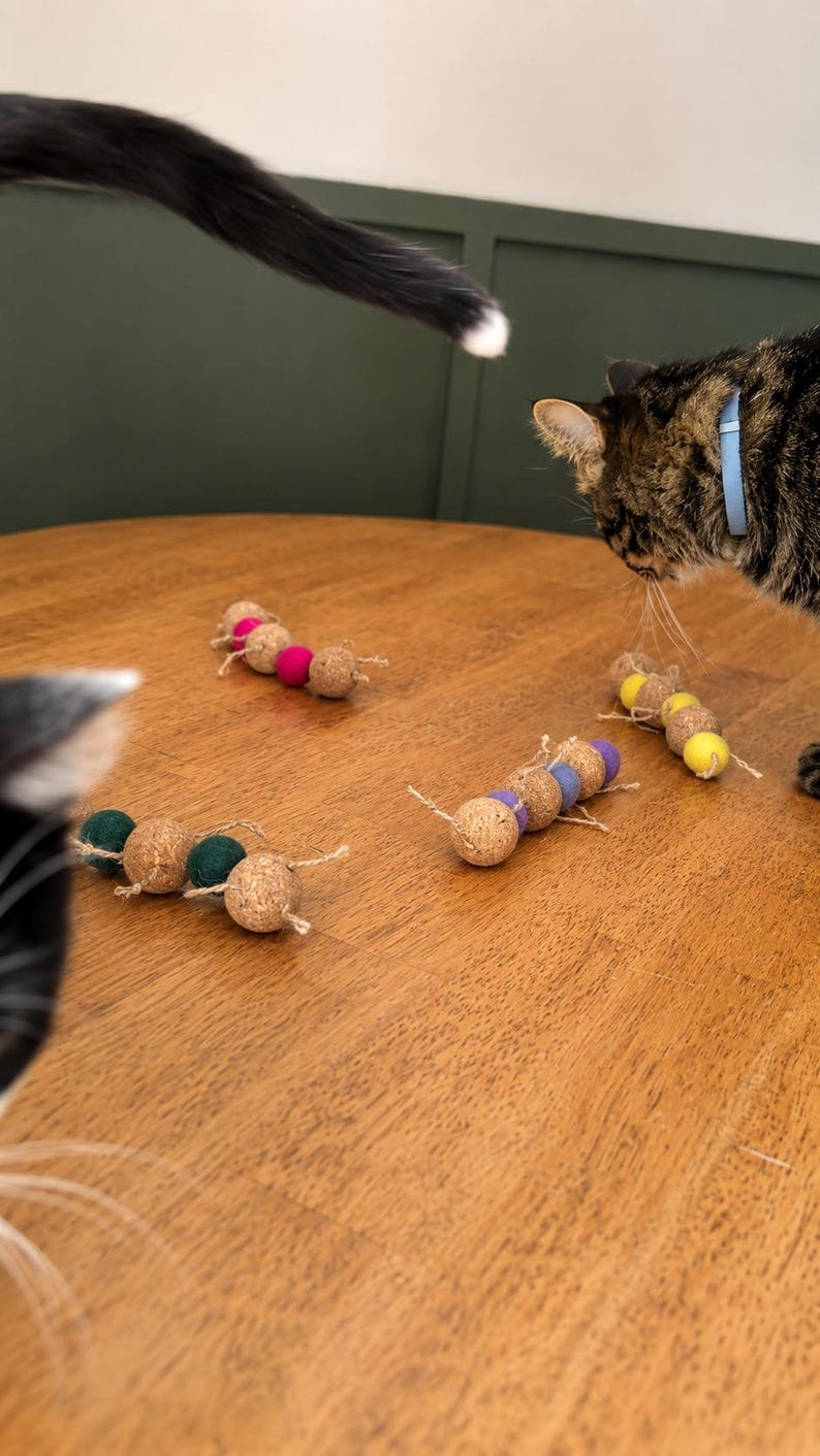 May include: Several cat toys made of cork and colorful felt balls are arranged on a wooden table. The toys are in a caterpillar-like design, with each segment a cork ball and a felt ball. A cat's tail and two cats are visible.