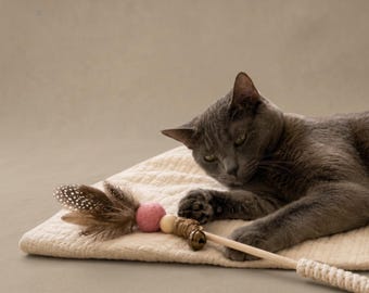 May include: A cat toy featuring a wooden wand, a small metal bell, a natural wooden bead, a pink felt ball, and brown spotted feathers. The toy is positioned on a textured cream-colored fabric surface next to a gray cat resting its paw on the wand.