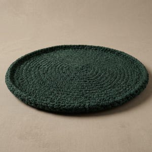 May include: A circular, dark forest green crochet mat featuring a dense, spiraling stitch pattern. The mat has a slightly raised, thick border edge and a flat, textured surface created by the interlocking yarn loops throughout the entire piece.