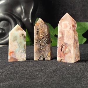 May include: Three gemstone towers in shades of pink, brown, and green stand on a dark gray surface. A black cat figurine is in the background. The crystals have a polished, pointed top and a rough, textured body.