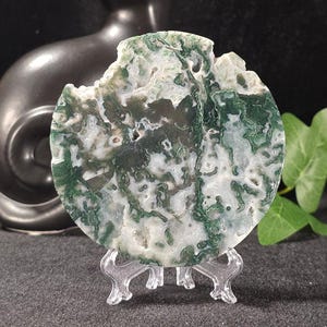 May include: A round, moss agate stone with a unique pattern of green and white swirls. The stone is displayed on a clear plastic stand, with a black cat statue in the background. The agate has a natural, organic shape.