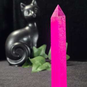 May include: A vibrant, hot pink crystal point stands tall, its surface textured. A black cat figurine and green leaves are in the background. The crystal is a striking decorative item.