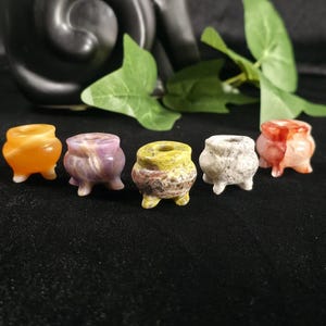 May include: Five small, carved stone cauldrons arranged in a row. The cauldrons are made of various minerals in orange, purple, yellow, white, and red hues. Each piece features a rounded body, a wide opening at the top, and three small feet.