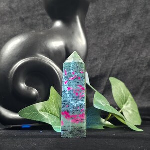 May include: A polished hexagonal crystal tower featuring a mottled green and black base with vibrant, irregular magenta-pink inclusions throughout the stone, standing upright with a pointed top and a smooth, reflective surface texture.
