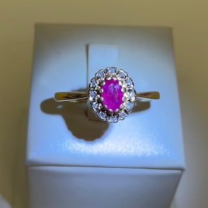 May include: A gold ring with an oval ruby gemstone, encircled by small diamonds. The ring's band is slender, highlighting the central stone. The ring is presented on a white surface. The ruby is a deep pink colour.