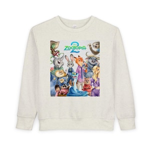 May include: A light beige sweatshirt featuring a colorful graphic of characters from the movie "Zootopia 2". The graphic includes the movie title and various animal characters in a watercolor style. The sweatshirt has long sleeves and a crew neck.
