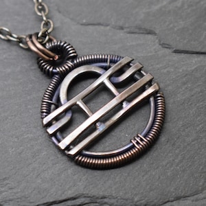 Könnte beinhalten: Ein runder Anhänger mit dem Waage-Sternzeichen-Symbol aus dunklem Metall, eingefasst in einen gewickelten Drahtrahmen. Das Design besteht aus horizontalen Stäben und einem Bogen, befestigt an einer dunklen Metallkette mit einer Drahtöse.