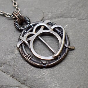May include: A silver pendant necklace with a circular design featuring a smaller circle inside. The pendant is made of wire and has a rustic, industrial look.