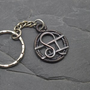 May include: A silver-toned keychain with a circular key ring and a decorative pendant. The pendant features a stylized design within a wire-wrapped circle, connected by a chain. The background is a dark, textured surface.