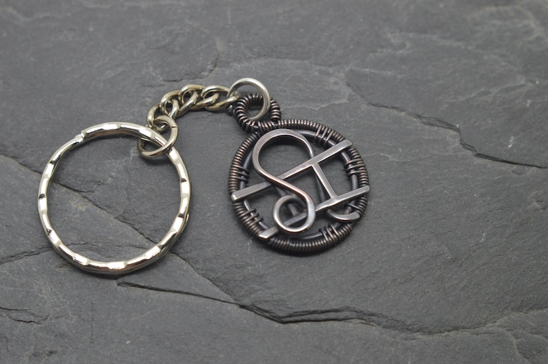 May include: A silver-toned keychain with a circular key ring and a decorative pendant. The pendant features a stylized design within a wire-wrapped circle, connected by a chain. The background is a dark, textured surface.