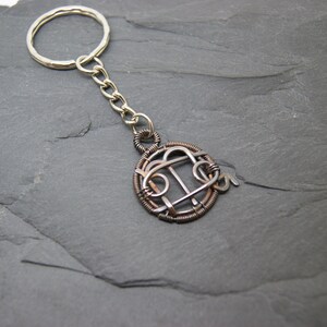 May include: A handmade keychain with a silver key ring and chain. The pendant is a circular design made of copper and silver wire, featuring an abstract pattern. The keychain is displayed on a dark gray, textured surface.
