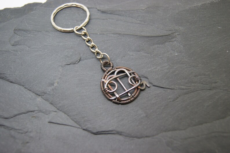 May include: A handmade keychain with a silver key ring and chain. The pendant is a circular design made of copper and silver wire, featuring an abstract pattern. The keychain is displayed on a dark gray, textured surface.