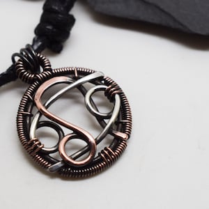 May include: A silver and copper wire wrapped pendant necklace featuring the astrological symbol for Leo. The pendant is round and hangs from a black cord.