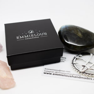 May include: A black box with the text "Emmielous Handmades" on the top. The box is surrounded by various crystals, including a large green labradorite, a pink rose quartz, and a purple amethyst. A silver charm with a delicate design is also visible.