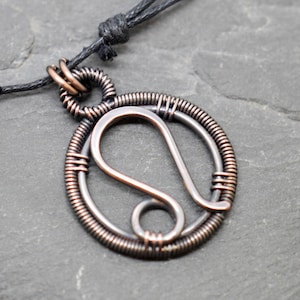 Op de afbeelding: Een koperen draadketting met een ronde hanger met het astrologische symbool voor Leeuw. De hanger is gemaakt van draad dat is gedraaid en gewikkeld om een ​​gedetailleerd ontwerp te creëren.