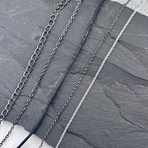May include: A collection of five distinct necklace cords and chains arranged diagonally. The set includes a thick dark metal link chain, a thin dark metal chain, a black cord, a thin dark metal chain, and a thin silver-coloured metal chain.