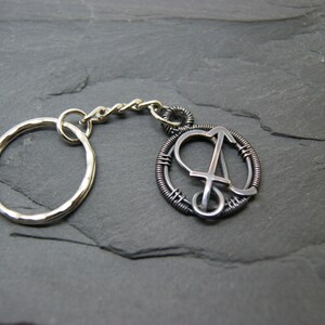 May include: Silver-toned keychain with a circular pendant featuring the letter "A" in a stylized design. The pendant is encircled by a wire-wrapped frame. The keychain includes a split ring and a chain.