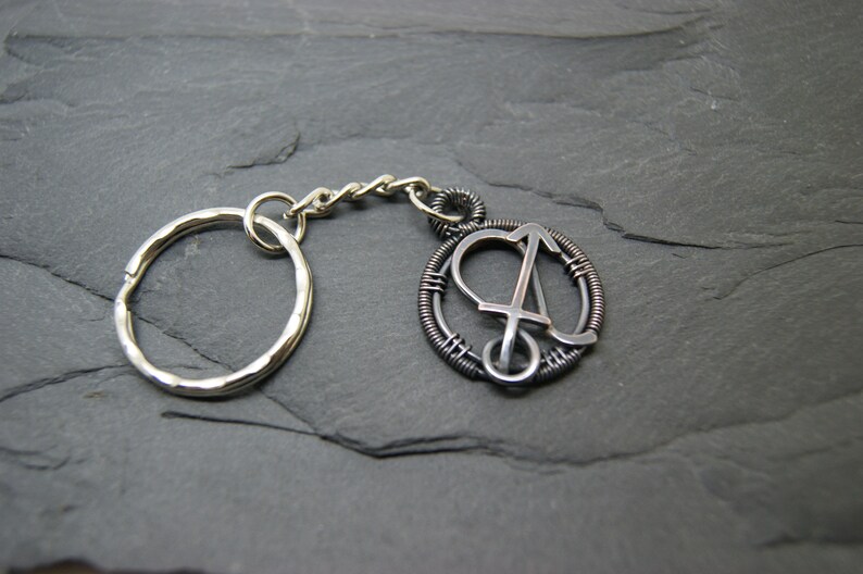 May include: Silver-toned keychain with a circular pendant featuring the letter "A" in a stylized design. The pendant is encircled by a wire-wrapped frame. The keychain includes a split ring and a chain.
