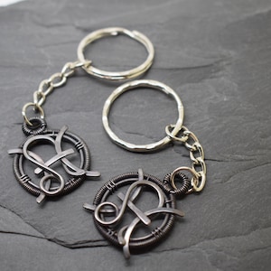 May include: Two handmade keychains with a dark metal wire design. Each keychain features a circular frame with an intertwined letter design, connected to a silver chain and key ring. The background is a dark gray, textured surface.