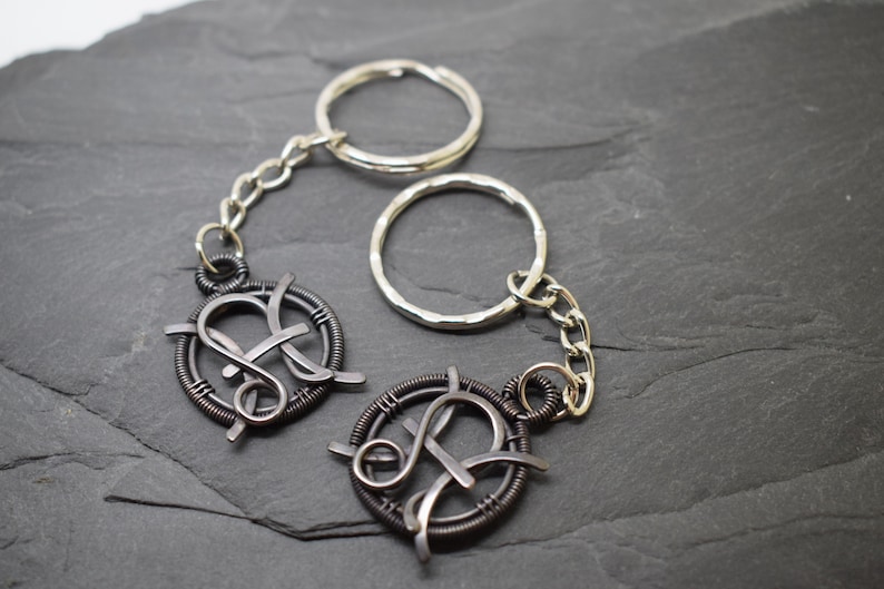 May include: Two handmade keychains with a dark metal wire design. Each keychain features a circular frame with an intertwined letter design, connected to a silver chain and key ring. The background is a dark gray, textured surface.