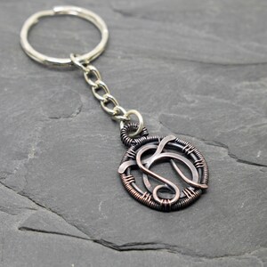 May include: A handcrafted keychain featuring a circular pendant with a wire-wrapped design. The pendant has a silver-colored chain and key ring. The pendant's design is a stylized letter 'S' in a copper and silver color scheme.