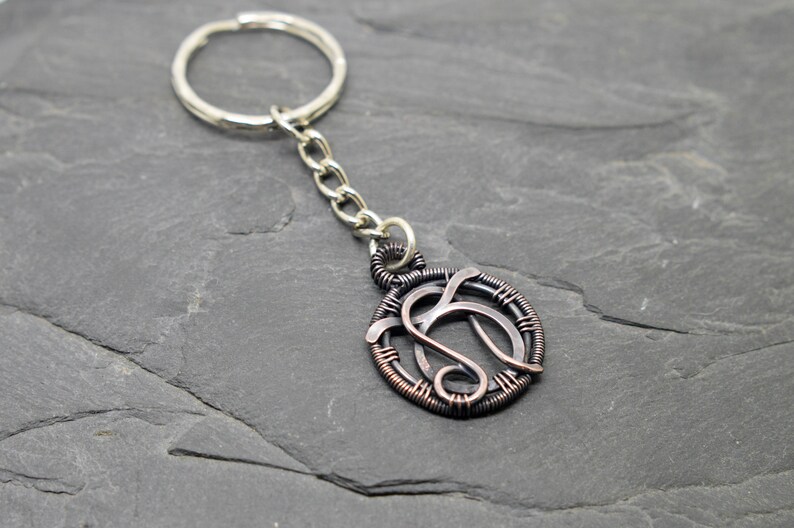 May include: A handcrafted keychain featuring a circular pendant with a wire-wrapped design. The pendant has a silver-colored chain and key ring. The pendant's design is a stylized letter 'S' in a copper and silver color scheme.