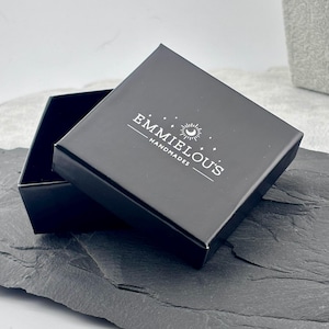 May include: A black gift box with the text "EMMIELOU'S HANDMADES" in white. The box is open, revealing a dark interior. The box is on a dark gray surface.