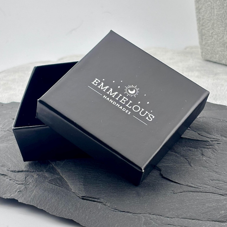 May include: A black gift box with the text "EMMIELOU'S HANDMADES" in white. The box is open, revealing a dark interior. The box is on a dark gray surface.