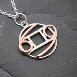 May include: A silver and rose gold necklace with a pendant shaped like the astrological sign Cancer. The pendant is made of two circles, one inside the other, with a square in the center. The outer circle is silver, and the inner circle and square are rose gold.