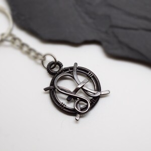 May include: A handcrafted, dark metal keychain featuring a circular design with an intertwined letter "K" in the center. The keychain includes a chain and a silver-toned key ring. The background is a white surface with a dark gray, textured accent.