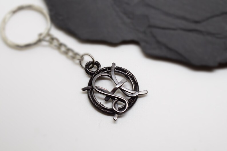 May include: A handcrafted, dark metal keychain featuring a circular design with an intertwined letter "K" in the center. The keychain includes a chain and a silver-toned key ring. The background is a white surface with a dark gray, textured accent.
