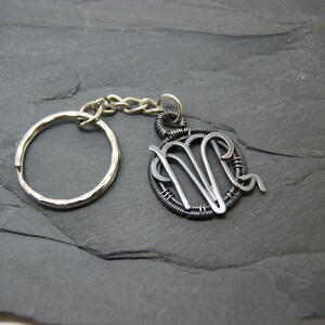 May include: A silver-toned keychain featuring a Virgo zodiac symbol pendant. The pendant is a wire-wrapped design with the Virgo symbol in the center. The keychain includes a split ring and a chain, all in silver tones.