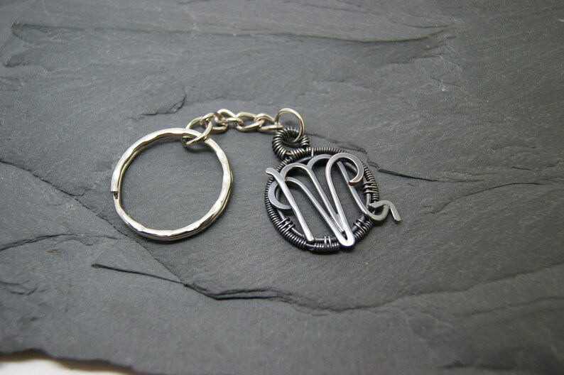 May include: A silver-toned keychain featuring a Virgo zodiac symbol pendant. The pendant is a wire-wrapped design with the Virgo symbol in the center. The keychain includes a split ring and a chain, all in silver tones.