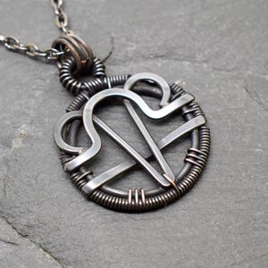May include: A dark metallic pendant featuring a stylised Libra zodiac symbol crafted from wire. The circular frame is wrapped in thin wire coils, and the piece hangs from a matching dark metal chain, showcasing a weathered, oxidised finish.