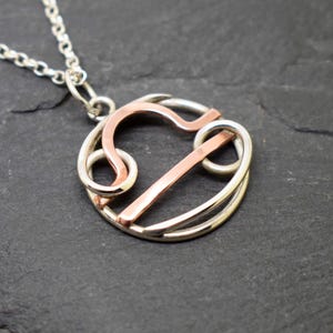 May include: A circular pendant necklace featuring the Libra zodiac symbol crafted from intertwined silver and copper wire. The pendant hangs from a delicate silver chain, showcasing a mixed-metal design with a polished finish and open wirework.