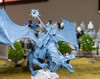 Może przedstawiać: Jasnoniebieska miniatura fantasy przedstawiająca smoka z jeźdźcem trzymającym laskę. Smok siedzi na skale z rozpostartymi skrzydłami. Miniatura jest bardzo szczegółowa i wydaje się być wykonana z plastiku.