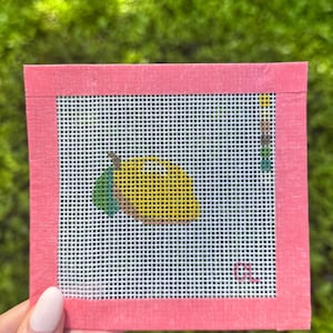 May include: A needlepoint canvas featuring a yellow lemon with green leaves and brown accents. The canvas is square, with a pink border, and a white grid background. The design is hand-stitched, showcasing a fruit theme.