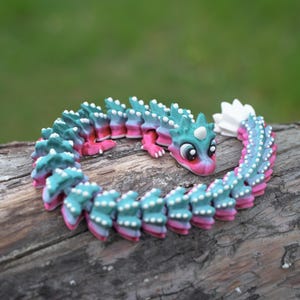 May include: A vibrant, articulated dragon toy in teal, pink, and blue, displayed on a wooden surface. The dragon features a detailed design with white accents and a friendly expression. A unique and whimsical toy.