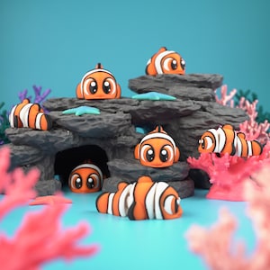 May include: A collection of orange and white clownfish figurines with black accents, arranged around a gray rock formation and coral. The scene is set against a blue background, with small blue starfish scattered around.