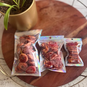 May include: Three clear, iridescent-edged bags filled with red-colored, dried fruit slices. The bags are arranged on a wooden serving tray with a gold-colored metal rim. A small potted plant is in the background.