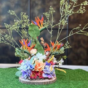 May include: A vibrant floral arrangement featuring orange, purple, and blue flowers, including bird of paradise blooms. The arrangement is set on a wooden base with a green moss-like surface, creating a natural aesthetic. Delicate green sprigs add height and texture.
