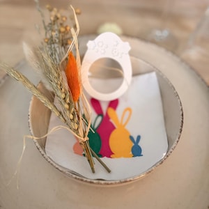 Può includere: Un piatto in ceramica beige con una ciotola più piccola contenente un tovagliolo e elementi decorativi. Il tovagliolo presenta sagome di coniglietti pasquali colorati. Un portatovagliolo bianco a forma di uovo e fiori secchi.