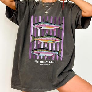 May include: A charcoal gray short-sleeve t-shirt featuring a graphic of three rainbow trout swimming horizontally over vertical purple brushstrokes. Below the fish, the text reads Fishers of Men and Matthew 4:19 in a clean white font.