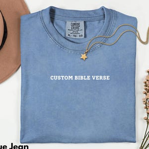 May include: A blue jean coloured t-shirt with the text "CUSTOM BIBLE VERSE" printed in white. A gold necklace with a star pendant is draped across the shirt. A brown hat is visible in the upper left corner.