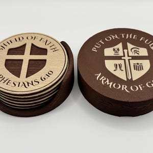 Op de afbeelding: Een set ronde bruine onderzetters met een houtnerftextuur, opgeborgen in een bijpassende houder. Eén onderzetter heeft een schildpictogram met de tekst SHIELD OF FAITH EPHESIANS 6:10. Het deksel toont een schild met harnaspictogrammen en PUT ON THE FULL ARMOR OF GOD.