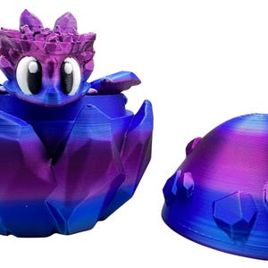 May include: A 3D-printed fantasy creature container in shades of blue, purple, and pink. The container is open, revealing a cartoon-like character with large white eyes. A matching lid is next to the container.
