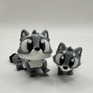 May include: Two 3D-printed raccoon figurines in varying sizes, featuring a layered gray and white texture. Both figures have large, dark, rounded eyes, pointed ears, and a small black nose, with the larger raccoon positioned on the left.