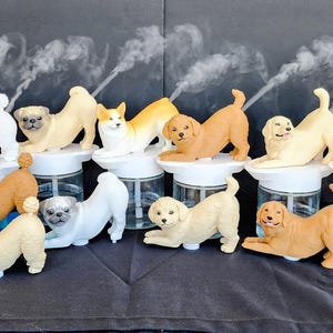 May include: A collection of small, portable humidifiers, each topped with a different dog or cat figurine in a stretching pose. The clear glass bases hold water, and each unit emits a thin stream of mist from the animal's back, showcasing various breeds.