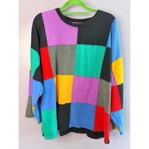 May include: A multi-coloured patchwork sweatshirt with long sleeves, featuring a mix of black, purple, red, green, yellow, and blue squares. The garment is displayed on a green hanger. The design is made up of large, square colour blocks.